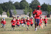 Toddlers running behind Sportball coach on soccer field.